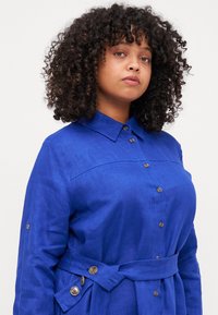 Abito camicia in lino blu con bottone frontale, colletto, vita con lacci e maniche arrotolate, caratterizzato da bottoni in tonalità dorata e un tessuto testurizzato.
