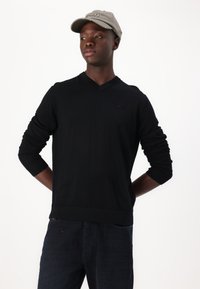 Maglione nero a V con maniche lunghe, realizzato in un materiale morbido. Presenta un piccolo logo sul petto. Indossato con jeans scuri.