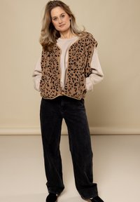 Bruine fleece vest met zwarte luipaardvlekken, gedragen over een lichtbeige top, gecombineerd met zwarte wijde jeans en zwarte schoenen. Neutrale achtergrond.