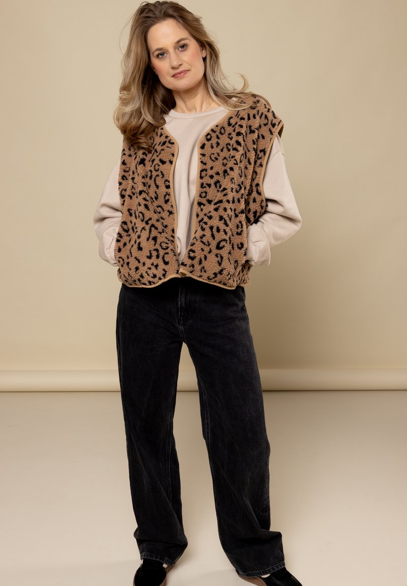 Bruine fleece vest met zwarte luipaardvlekken, gedragen over een lichtbeige top, gecombineerd met zwarte wijde jeans en zwarte schoenen. Neutrale achtergrond.