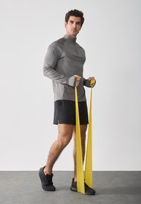 Mannen sportoutfit met een grijze lange mouwen rits-top, zwarte shorts en zwarte sneakers. Gele weerstandsbanden in de handen.