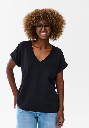 Kobieta o kręconych włosach i w okularach, uśmiechnięta, ubrana w czarną koszulkę z dekoltem w serek i jasnoniebieskie dżinsy, stojąca na białym tle.