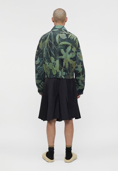 Άτομο με ξυρισμένο κεφάλι που φοράει πράσινο και μαύρο φλοράλ μπουφάν, μαύρη πλισέ φούστα, μαύρες κάλτσες και μπεζ παντόφλες, στεκόμενο με την πλάτη στραμμένη.