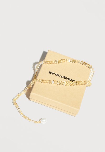 Collier délicat en perles dorées avec un pendentif en grosse perle blanche posé sur une boîte à bijoux beige Loren Stewart.