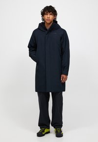 Marine waterdichte jas met capuchon, voorzien van een sluiting aan de voorkant en zakken, gecombineerd met donkere broek en zwart schoenen met gele accenten.