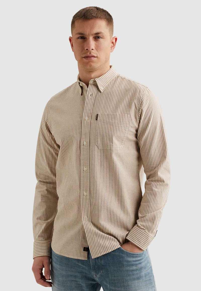 Mladý muž oblečený v béžovo-bielej zvislo pruhovanej košeli s náprsným vreckom a modrých džínsoch, stojaci s jednou rukou v vrecku.