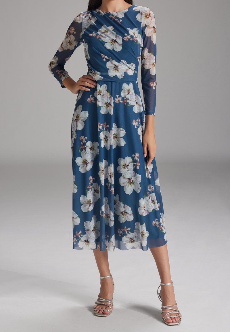 Femme portant une robe midi bleue à imprimé floral blanc et manches longues transparentes, assortie à des sandales argentées à talons hauts et à brides.