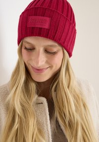 Rode gebreide beanie met ribbelstructuur en een rechthoekige leren patch met de tekst "VIND JE INNERLIJKE VREDE." Model draagt een beige jas.