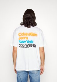 Biała bawełniana koszulka z kolorowym napisem na plecach: „Calvin Klein Jeans” w kolorze pomarańczowym, „New York” w kolorze niebieskim, wraz z dodatkowymi czarnymi napisami.