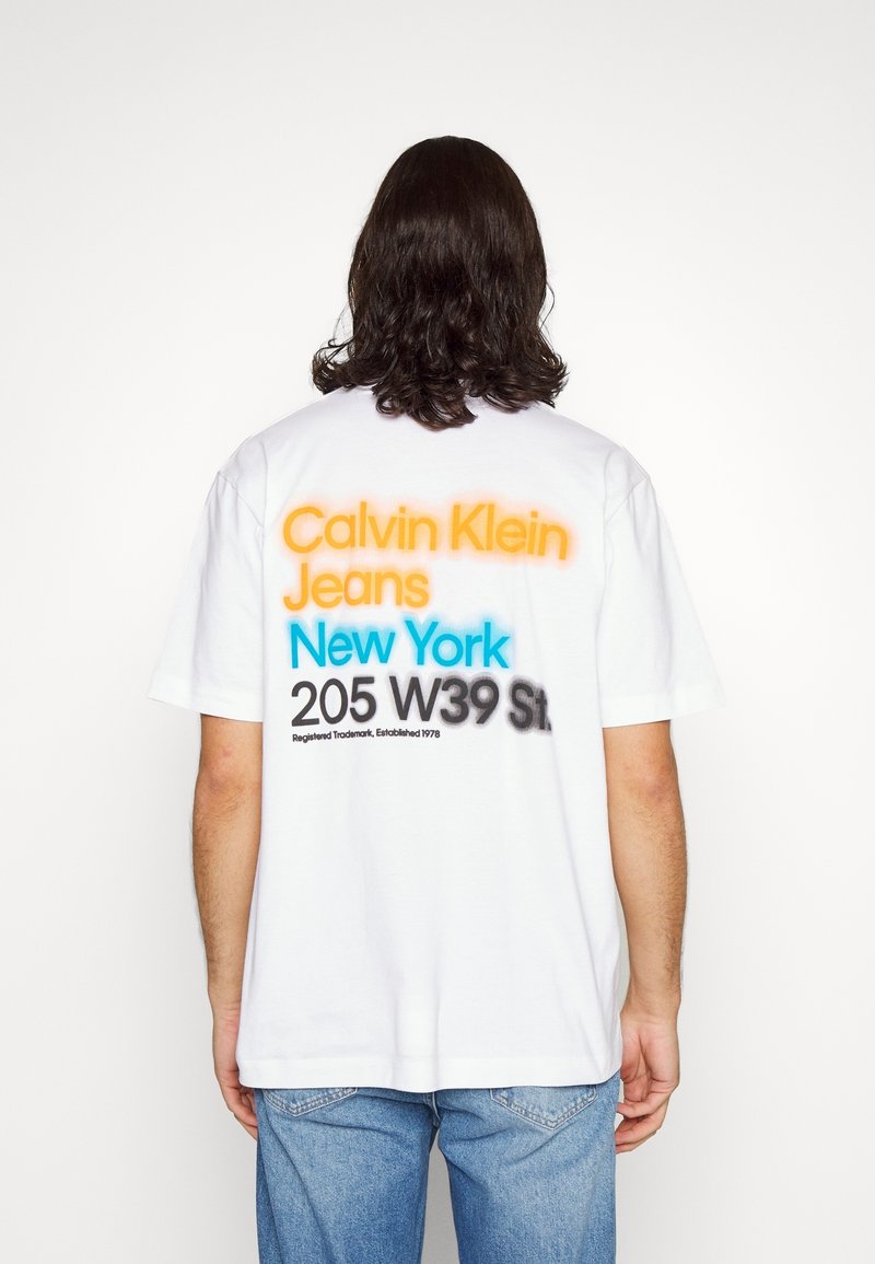 Biała bawełniana koszulka z kolorowym napisem na plecach: „Calvin Klein Jeans” w kolorze pomarańczowym, „New York” w kolorze niebieskim, wraz z dodatkowymi czarnymi napisami.