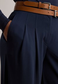 Asmuo vilkintis tamsiai mėlynas klostuotas kelnes su rudu odiniu diržu su dviem sagtimis, ranka ilsisi kišenėje, prieš paprastą foną.