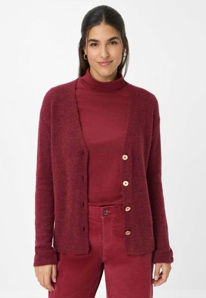 Burgunderfarbener Cardigan mit weicher Textur, ausgestattet mit einem V-Ausschnitt, sechs Knöpfen und langen Ärmeln. Über einem passenden Rollkragenpullover getragen.