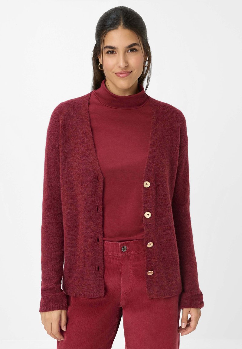 Burgunderfarbener Cardigan mit weicher Textur, ausgestattet mit einem V-Ausschnitt, sechs Knöpfen und langen Ärmeln. Über einem passenden Rollkragenpullover getragen.