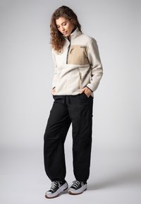 Fleece-Pullover in Creme mit einer hellbraunen Brusttasche, halbem Reißverschluss und kontrastierendem marineblauem Saum, kombiniert mit schwarzen Cargo-Hosen und Turnschuhen.