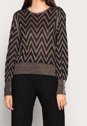 Femme portant un pull à manches longues noir et or à motif chevron pailleté, avec des poignets boutonnés et un pantalon noir.