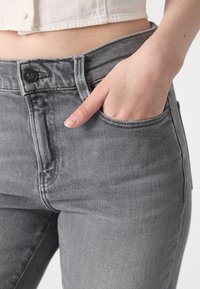 Los pantalones vaqueros de mezclilla gris presentan un cierre de botón y cremallera, bolsillos laterales y una silueta ajustada, con costuras visibles y una textura suave.