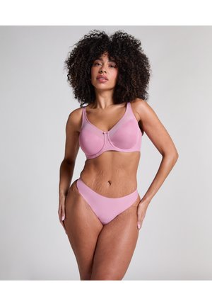 Frau mit lockigem Haar, die selbstbewusst steht und einen passenden hellrosa BH und Unterwäsche vor einem einfarbigen Hintergrund trägt.