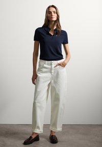 Polo shirt navy con maniche corte, abbinato a jeans bianchi a vita alta e a gamba larga. Mocassini scuri completano l'outfit. Sfondo semplice.