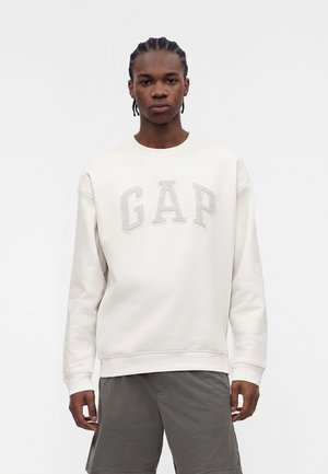 Jonge man met gevlochten haar, gekleed in een witte GAP-trui en grijze short, staand tegen een effen witte achtergrond.