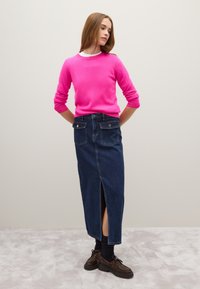 Suéter de punto fucsia con mangas tres cuartos, combinado con una falda larga de mezclilla oscura con una abertura en el frente y dos bolsillos traseros. Zapatos marrones.