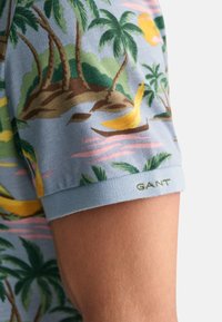 Košile s krátkým rukávem z bavlny s modrým pozadím, na kterém jsou tropické palm trees, banány a zelené logo GANT na manžetě.