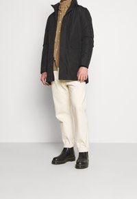 Veste noire imperméable avec capuche, fermée par des boutons, et poches latérales, portée sur une chemise marron et un pantalon crème, associée à des bottes noires.