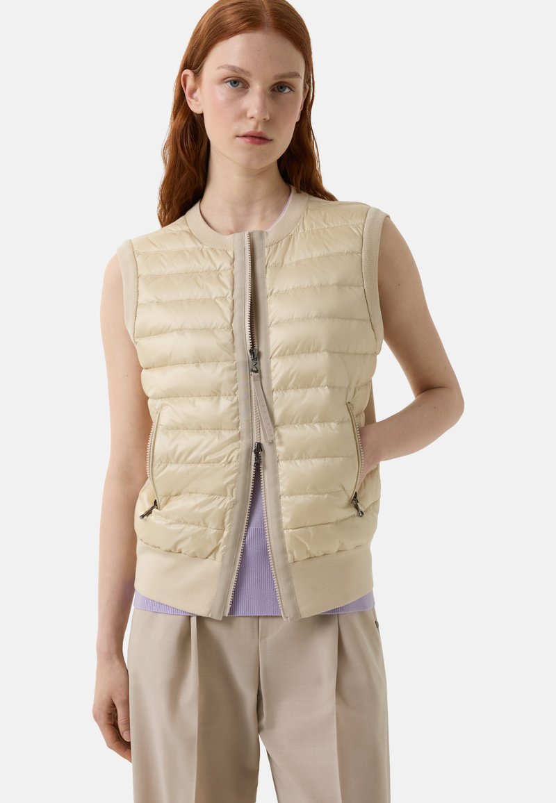 Ärmelleste beige Pufferweste mit einer taillierten Passform, horizontal gestepptem Muster, zwei Seitentaschen und einem Frontreißverschluss. Getragen über einem hellvioletten Oberteil.