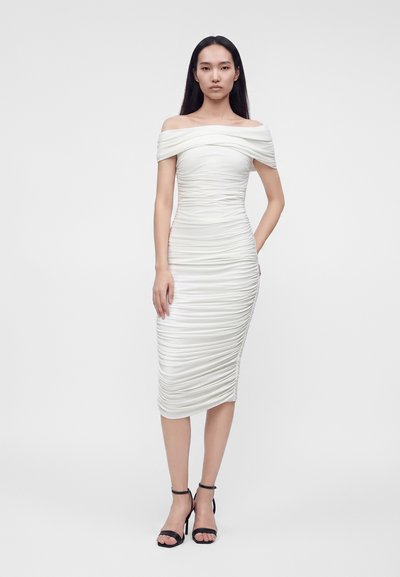 Kvinde iført en tætsiddende hvid off-shoulder midi-kjole med rynker og sorte højhælte sandaler, stående mod en ensfarvet baggrund.