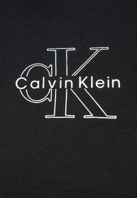 Μαύρο ύφασμα με μεγάλες λευκές περιγράμμενες αρχικές "C" και "K" που αλληλοεπικαλύπτονται, με το κείμενο "Calvin Klein" σε πεζά γράμματα τοποθετημένο στο κέντρο πάνω από τα γράμματα.