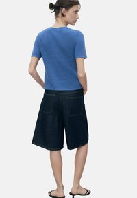 Haut en tricot côtelé bleu à manches courtes associé à un short en denim foncé avec poches arrière et coupe décontractée. Modèle portant des sandales noires.