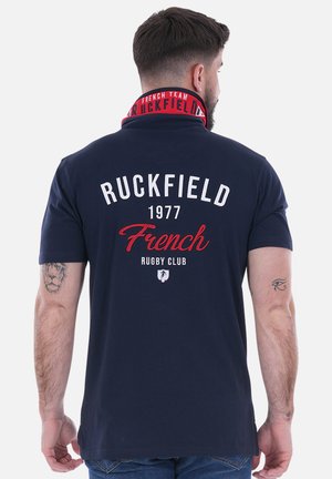Polo shirt marine avec un col rouge et blanc. Comprend "RUCKFIELD" et "French Rugby Club" imprimés au dos dans des couleurs contrastées.