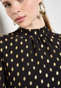 Schwarze Bluse mit hohem Kragen, gestaltet mit goldenen, metallischen Punktmustern und feinen Falten am Kragen für zusätzliche Textur.