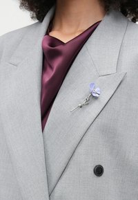 Harmaa bleiseri, jossa on teksturoitu kangas, yhdistettynä syvään burgundy-satiinipaitaan. Siinä on hopeinen kukkabroshi, jossa on laventelivärinen kukkasomistus.