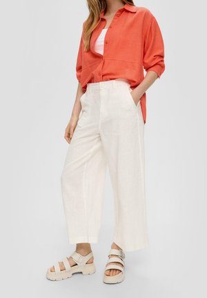 Frau trägt weite weiße Hose, orangefarbenes Hemd über weißem Top und beige klobige Riemchensandalen, steht mit einer Hand in der Tasche.