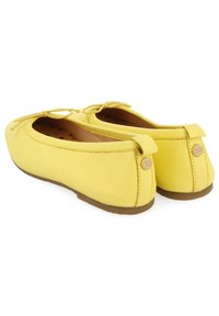 Zapatos planos amarillos con un acabado de tela suave, punta redonda, moño decorativo y un pequeño detalle de logo en la parte trasera. Suelas de goma marrón.