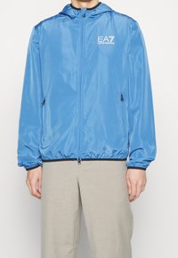 Ljusblå zip-jacka med huva. Tillverkad av blankt tyg, har elastiska muddar och midja. EA7-logotyp på bröstet.