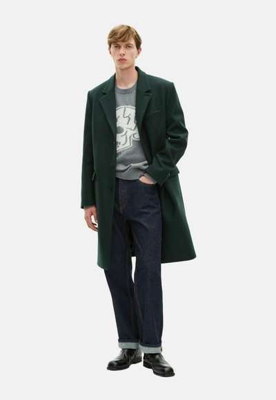 Abrigo de lana verde oscuro sobre un suéter gris claro con un diseño de calavera, combinado con jeans de mezclilla oscuros y botas negras hasta el tobillo.