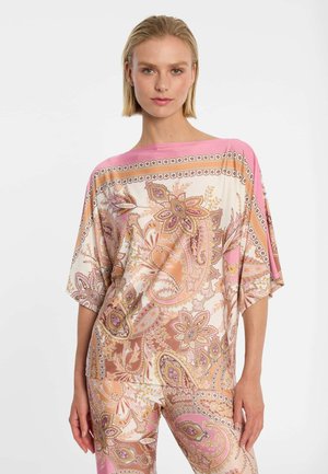 Frau trägt ein lockeres Oberteil mit Paisley-Muster in Pink, Beige und Weiß sowie passende Hosen, steht vor einem schlichten weißen Hintergrund.