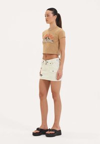 Afskåret beige t-shirt med grafisk print, parret med en hvid denim mini-skjørt med sidelommer og en sort blomstret detalje, samt sorte platform sandals.