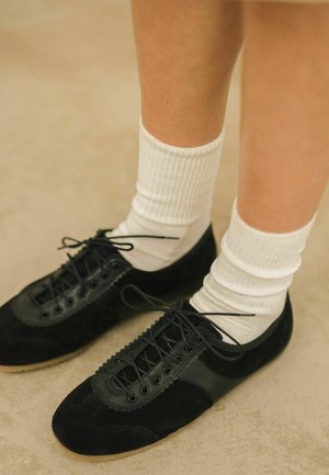 Scarpe in suede nere con accenti in pelle e suole in gomma, abbinate a calze bianche a coste, mostrate su un terreno chiaro e texturizzato.