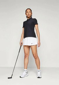 Μαύρο πόλο μπλουζάκι, λευκή πτυχωτή φούστα γκολφ, λευκά αθλητικά παπούτσια. Το μοντέλο κρατάει μια βέργα γκολφ, με φόντο έναν απλό γκρι τοίχο.