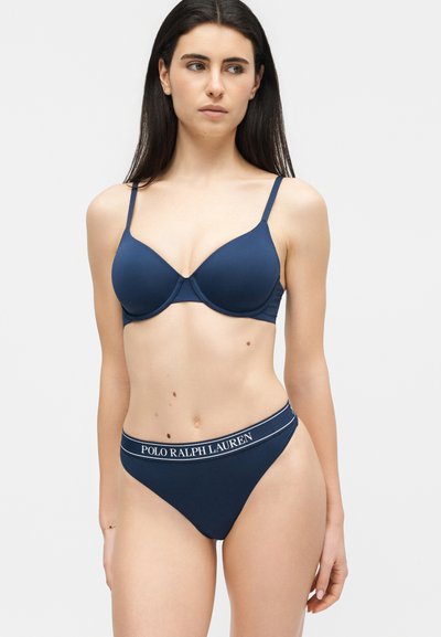 Γυναίκα με μακριά σκούρα μαλλιά που φοράει σουτιέν σε σκούρο μπλε και ταιριαστό σλιπ με το λογότυπο Polo Ralph Lauren στη μέση.