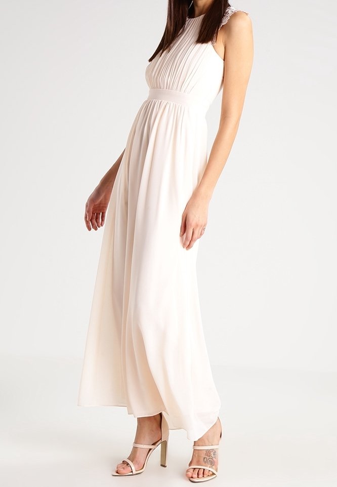 Longue robe fluide crème avec un corsage plissé et des accents en dentelle aux épaules. Portée avec des sandales argentées à talons hauts. Tissu lisse et léger.