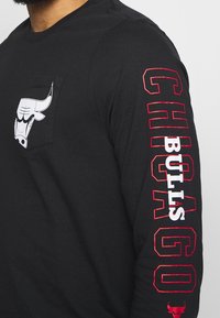 Czarna koszula z długim rękawem z przednią kieszenią i białym logo byka. Na rękawie znajdują się napis "CHICAGO" oraz "BULLS" w czerwonym i białym kolorze.