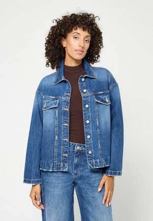 Frau mit lockigem Haar, die eine blaue Jeansjacke und Jeans über einem braunen Langarmshirt trägt, steht vor einem einfarbigen Hintergrund.