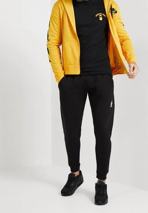 Mand iført en gul lynlås hoodie, sort grafisk t-shirt, sorte joggerbukser og sorte sneakers, står på en ensartet baggrund.