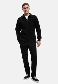 Suéter negro con cremallera de cuarto y cuello, con un bolsillo en el pecho, combinado con pantalones negros y zapatillas negras con detalles en blanco.
