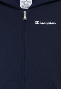 Tamnoplava zip-up dukserica izrađena od mekanog materijala, s bijelim Champion logotipom na lijevoj strani prsnog koša i glatkim patentnim zatvaračem sprijeda.