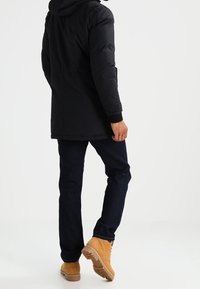 Svart isolerad jacka med huva, bärs över mörka jeans och beige stövlar. Har en lång skärning och ribbade manschetter. Enfärgad utan mönster.