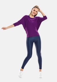 Violetti ylisuuri pusero, jossa on kolme neljäsosaa hihat, yhdistettynä kiiltaviin tummansinisiin leggingsseihin ja valkoisiin lenkkikenkiin. Silkkinen kangas, rento malli ja elävä väri.
